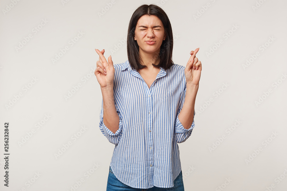 Brunette girl keeping her fingers crossed and eyes tightly closed while making a wish. Nice pretty girl sincerely believing in her luck, praying for close person,wanting some present or to win a prize Brunette girl keeping her fingers crossed and eyes tightly closed while making a wish. Nice pretty girl sincerely believing in her luck, praying for close person,wanting some present or to win a prize