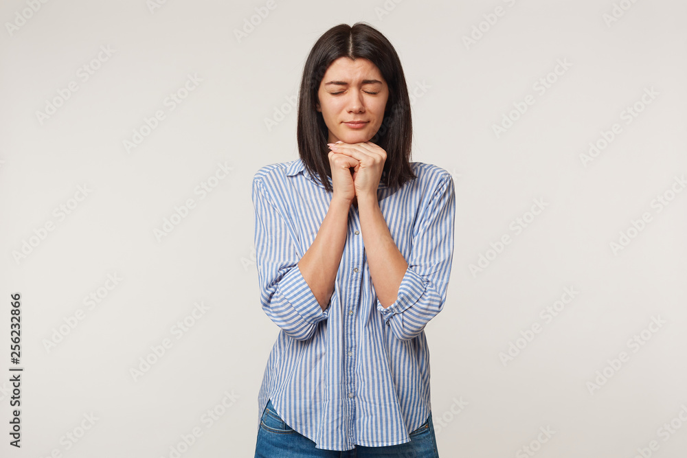 Girl folded hands in wishing gesture and eyes tightly closed while making a wish. Woman praying for close person, says thanks to God for help, wanting some present, hopes for luck, over white wall Girl folded hands in wishing gesture and eyes tightly closed while making a wish. Woman praying for close person, says thanks to God for help, wanting some present, hopes for luck, over white wall