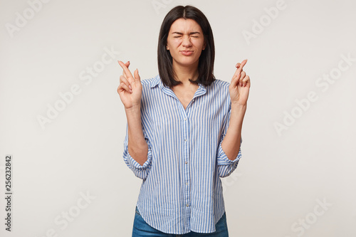 Brunette girl keeping her fingers crossed and eyes tightly closed while making a wish. Nice pretty girl sincerely believing in her luck, praying for close person,wanting some present or to win a prize