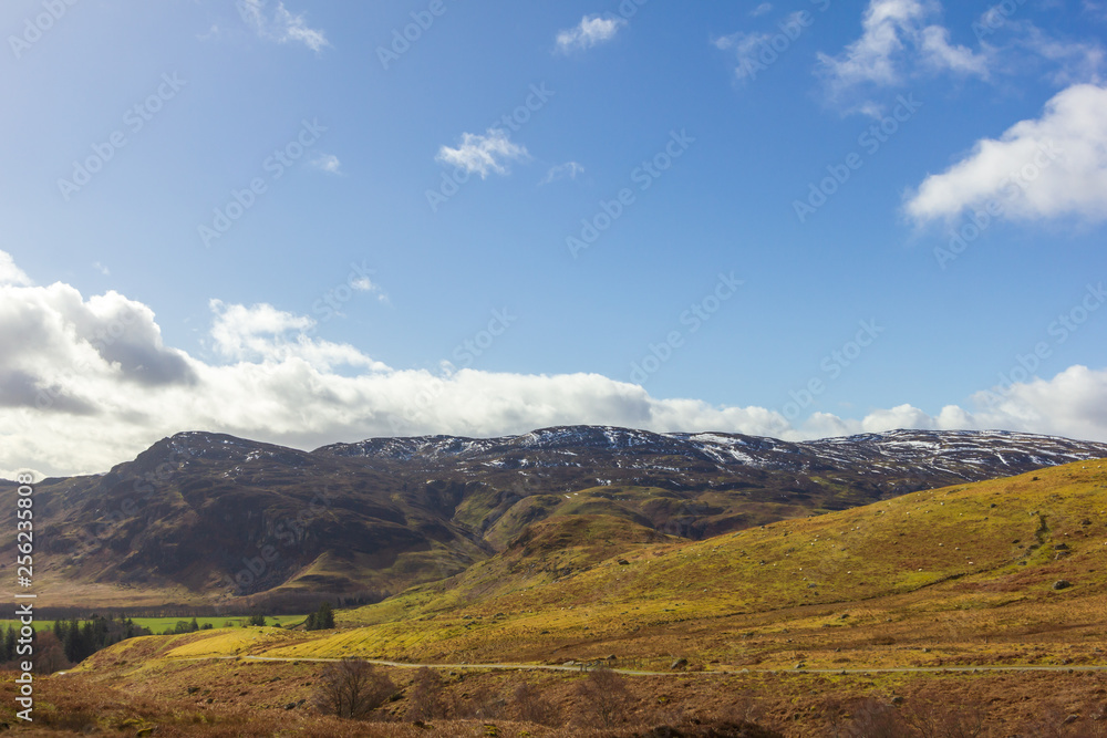 A view of mountain range with grassy green slopes and trail path under a majestic blue sky and white clouds