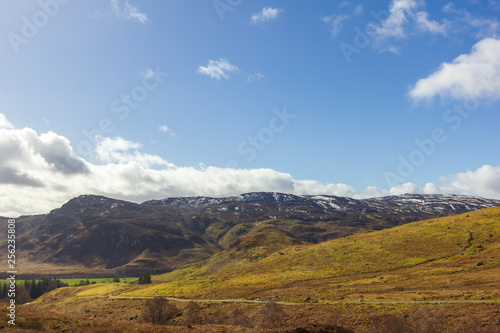 A view of mountain range with grassy green slopes and trail path under a majestic blue sky and white clouds © Dolwolfian