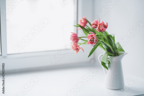 Pink peony tulips in a white jug stand on the window.
