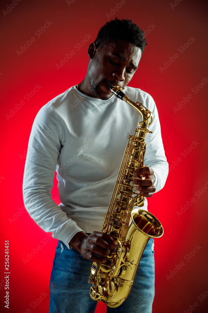 Naklejka premium African American handsome jazz musician playing the saxophone in the studio on a neon background. Music concept. Young joyful attractive guy improvising. Close-up retro portrait.