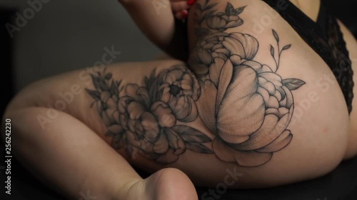 young woman in red bra and black underpants shows large peony tattoo on left thigh sitting in tattoo studio