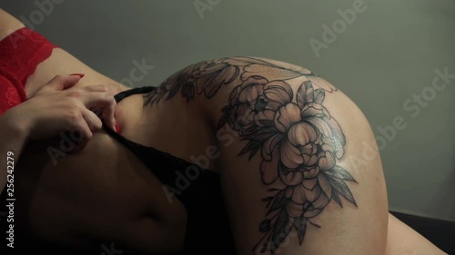 closeup young slim woman with red manicure and bra shows flower tattoo on left thigh lying in fashion studio