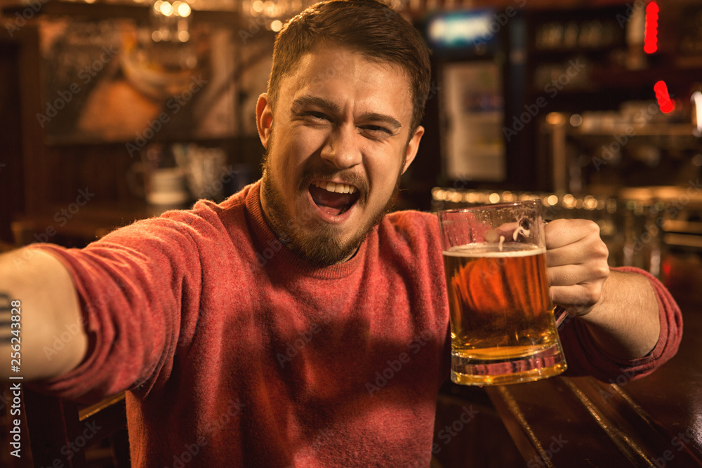 Cheerful handsome young man taking a selfie while drinking beer at the bar