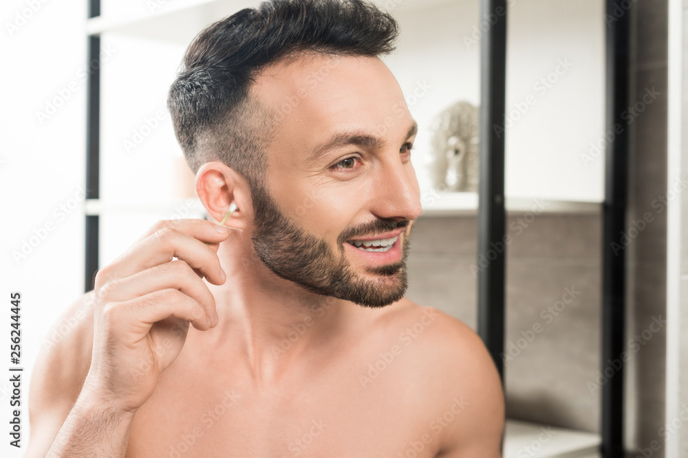 Fototapeta premium cheerful shirtless man cleaning ear while looking at mirror in bathroom