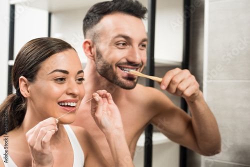 bearded boyfriend brushing teeth near girlfriend using dental floss in bathroom