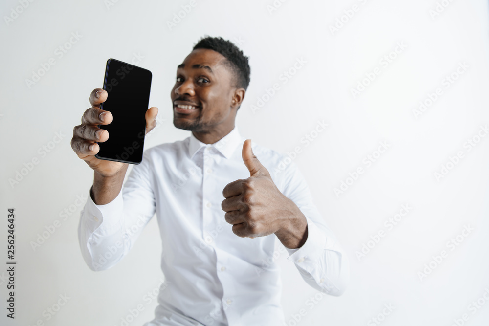 Indoor portrait of attractive young black african man isolated on grey background, holding blank smartphone, smiling at camera, showing screen, feeling happy and surprised. Human emotions, facial Indoor portrait of attractive young black african man isolated on grey background, holding blank smartphone, smiling at camera, showing screen, feeling happy and surprised. Human emotions, facial