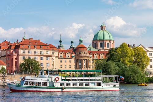 Sunny summer day on Vltava river with touristic boat in Prague. Cityscape with famous red roofs and green domes of baroque Church of St. Francis of Assisi and St. Salvator Church.