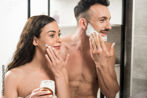handsome bearded man applying shaving foam on face near attractive girlfriend in bathroom