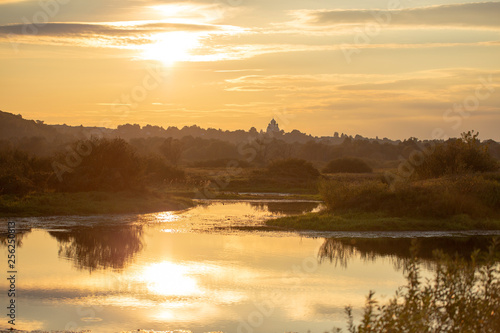 Amazing golden sunset rural landscape of sunny golden river and woods and old church silhouette in distance shooted during sunset time in autumn. Horizontal color photography.