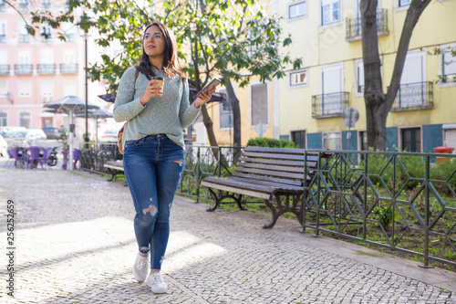 Content woman walking in city with smartphone and plastic cup