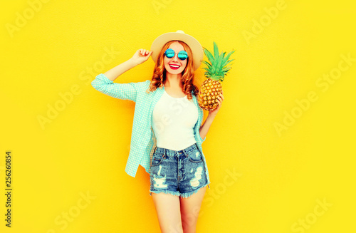 Happy smiling woman with pineapple having fun in summer straw hat, sunglasses, shorts on colorful yellow background