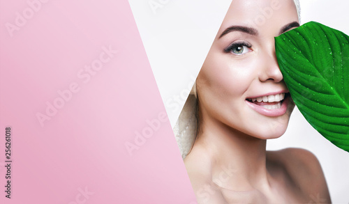 Portrait of young beautiful woman with healthy glow perfect smooth skin holds green tropical leaf, look into the hole of colored pink paper. Model with natural nude make up. Fashion, beauty, skincare.
