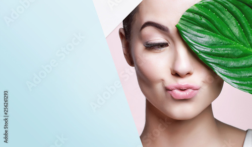Fototapeta Portret młoda piękna kobieta z zdrową poświatą perfect gładka skóra trzyma zielonego tropikalnego liść, patrzeje w dziurę kolorowy papier. Model z naturalnym makijażem nago. Moda, uroda, pielęgnacja skóry.