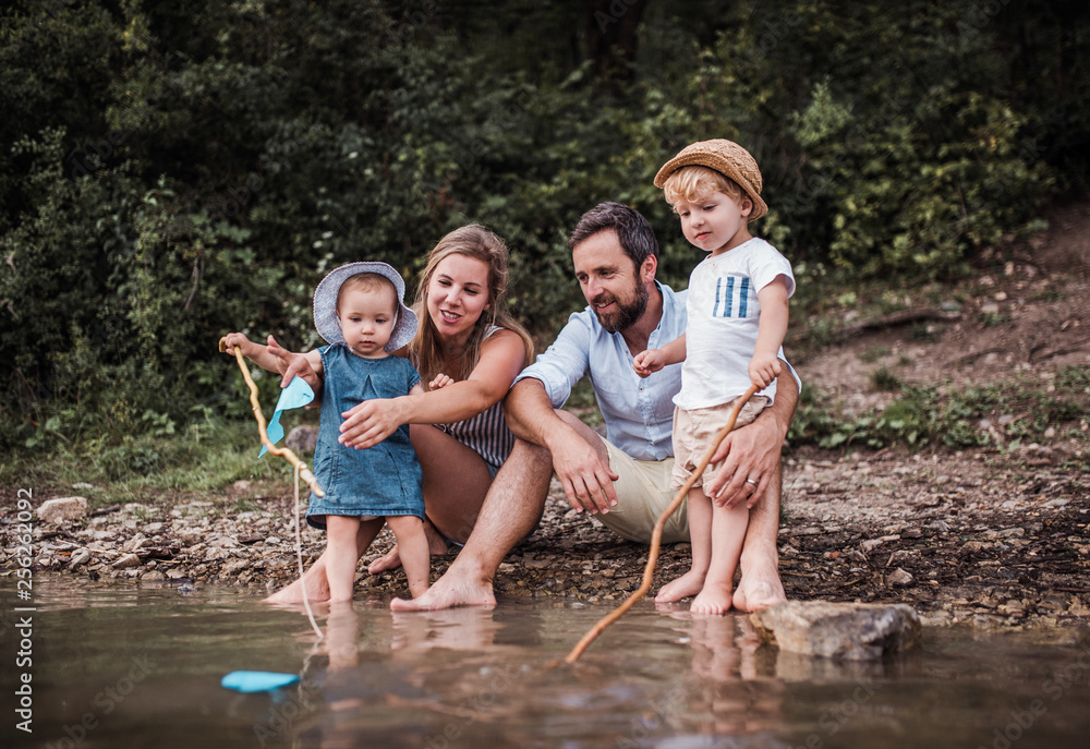 A young family with two toddler children outdoors by the river in summer. - obrazy, fototapety, plakaty 