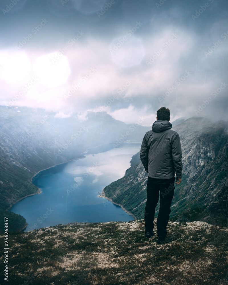 Junger Mann steht auf Gipfel über Fjord in Norwegen. Dramatischer Himmel und leichter Regen. Abenteuer in den Bergen. Blauer Fluss im Tal