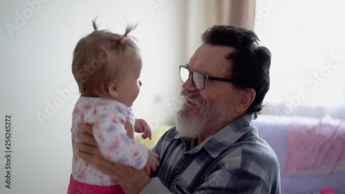 Grandfather with a beard, wearing glasses, playing with a little adorable granddaughter. He shakes her, they smile at each other, she plays with him. photo