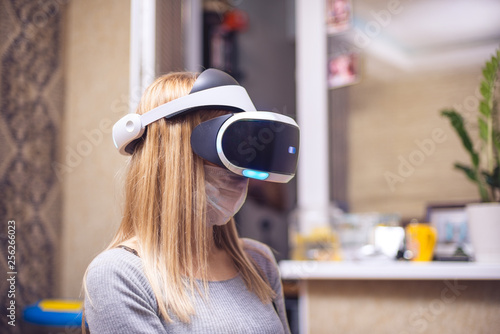 A young blond caucasian sick girl covers her face and wears a Virtual Reality Headset enjoys virtual reality in her apartment. She get pleasure from life, despite cold and feeling sick. Shallow DOF photo