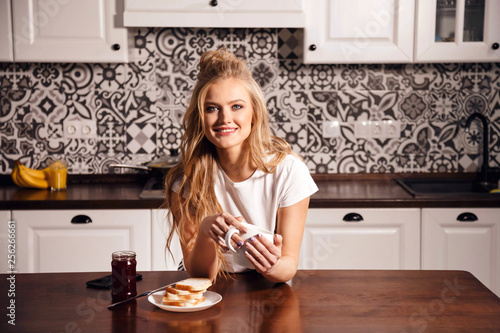 Adorable blonde girl in pajama enjoying hot coffee in modern light white kitchen interior