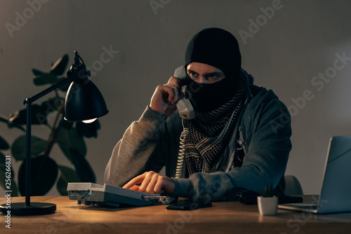 Serious criminal in black mask talking on telephone in room photo