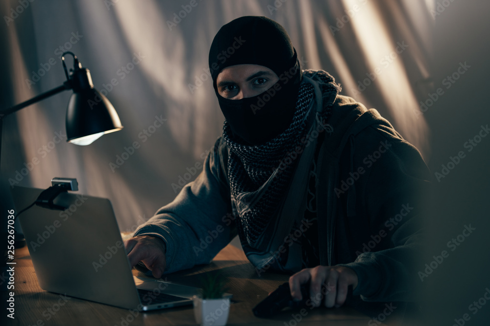 Terrorist in mask with gun using laptop in dark room Terrorist in mask with gun using laptop in dark room