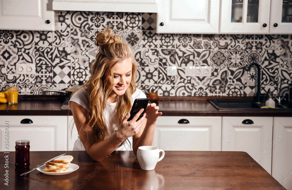 Image of blonde girl in pajama holding smartphone and cup of coffee in morning in kitchen Image of blonde girl in pajama holding smartphone and cup of coffee in morning in kitchen