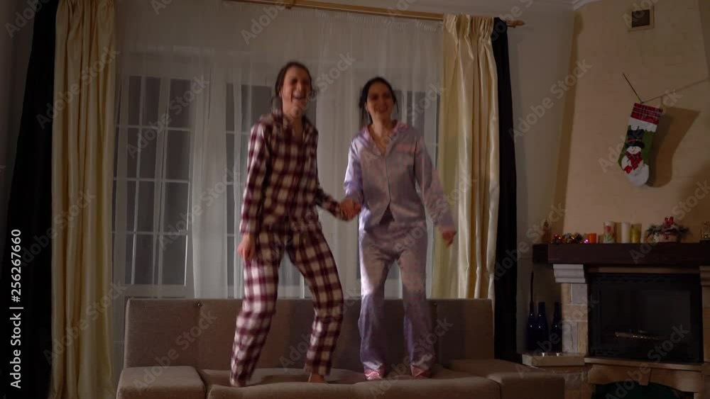 Two happy sisters twins in pajams jumping on the couch in a cozy living room and having fun like in childhood. Relationship sisters. Slow motion. Two happy sisters twins in pajams jumping on the couch in a cozy living room and having fun like in childhood. Relationship sisters. Slow motion.