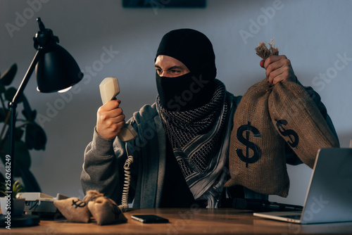 Fotografie Aggressive terrorist in mask holding money bags and looking at handset