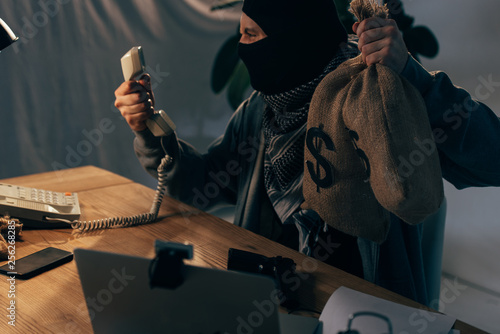 Angry terrorist in black mask holding money bags and looking at handset photo