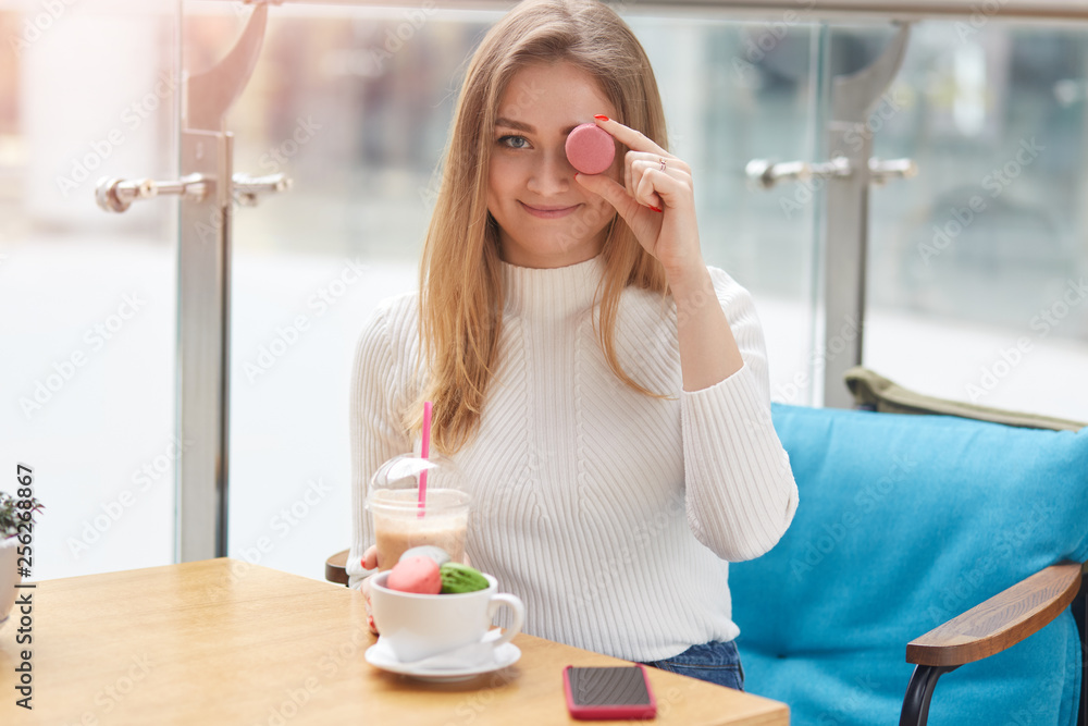 Young pretty lady in white shirt sits at table on blue sofa and covers her eye with pink macaroon while joyfully looking at camera in cafe. Attractive blonde female drinks milkshake. People concept. Young pretty lady in white shirt sits at table on blue sofa and covers her eye with pink macaroon while joyfully looking at camera in cafe. Attractive blonde female drinks milkshake. People concept.