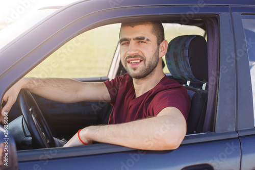 Handsome young dark haired male with stubble dressesd maroon casual t shirt, poses in black car, looks at camera trough window, enjoys his journey, being glad to drive. People, transportation concept. © sementsova321