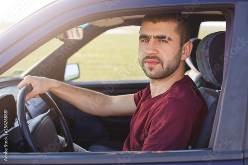 Portrait of attractive unshaved man with eyebrow strip, holds hand on steering wheel, drives black car, looks at camera, dressed casual red t shirt, has serious facial expressions. Traveling concept. photo