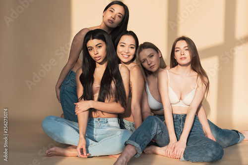 five attractive multicultural girls in blue jeans and bras smiling while looking at camera