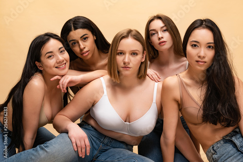 five pretty multicultural girls sitting and looking at camera isolated on beige, body positivity concept