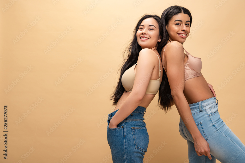 Fototapeta premium two multicultural happy girls in blue jeans and bra standing back to back and looking at camera isolated on beige, body positivity concept