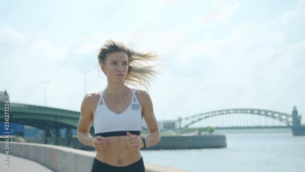 Medium shot of determined female athlete in white sports bra running along embankment near river on summer day in slow motion Medium shot of determined female athlete in white sports bra running along embankment near river on summer day in slow motion