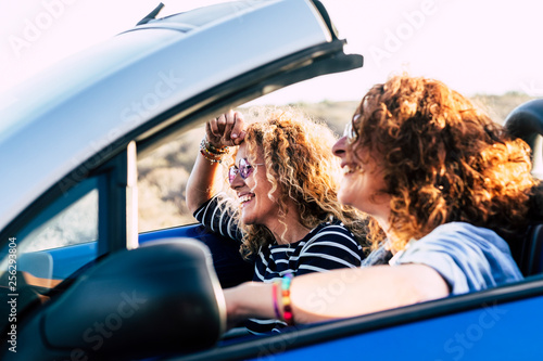 Two curly beautiful women friends drive and travel together on a convertible blue car having fun - outdoor happy leisure activity for cheerful people under the sun of summer - focus on second girl © simona