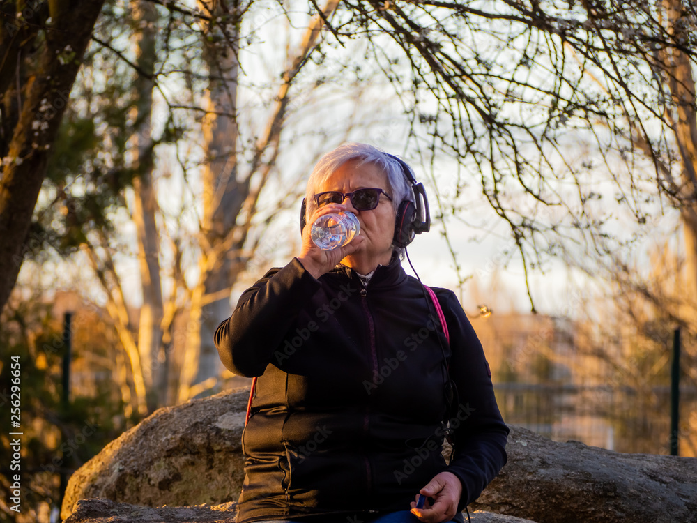 A senior woman with white hair listening to music with headphones and drinking water from a plastic bottle