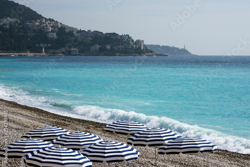 Striped blue and white umbrellas on a pebble beach on the Promenade des Anglais in Nice, France, await guests. Blue waves roll ashore on a sunny day. A cozy place for rest and relaxation.