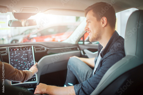 Young couple buying first electric car in the showroom. Attractive glad man looking at his girlfriend while she chooising a way on electronic dashboard in modern electric hybrid vehicle before test