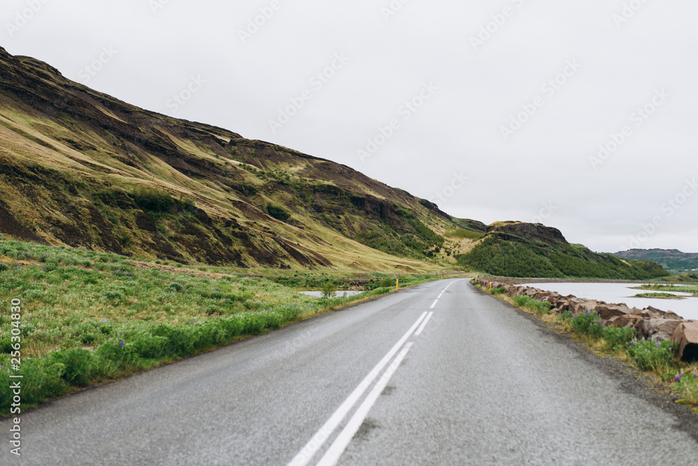 The road between the mountains and the hills. Asphalt road in Iceland. The road is in the field. The road between the mountains and the hills. Asphalt road in Iceland. The road is in the field.