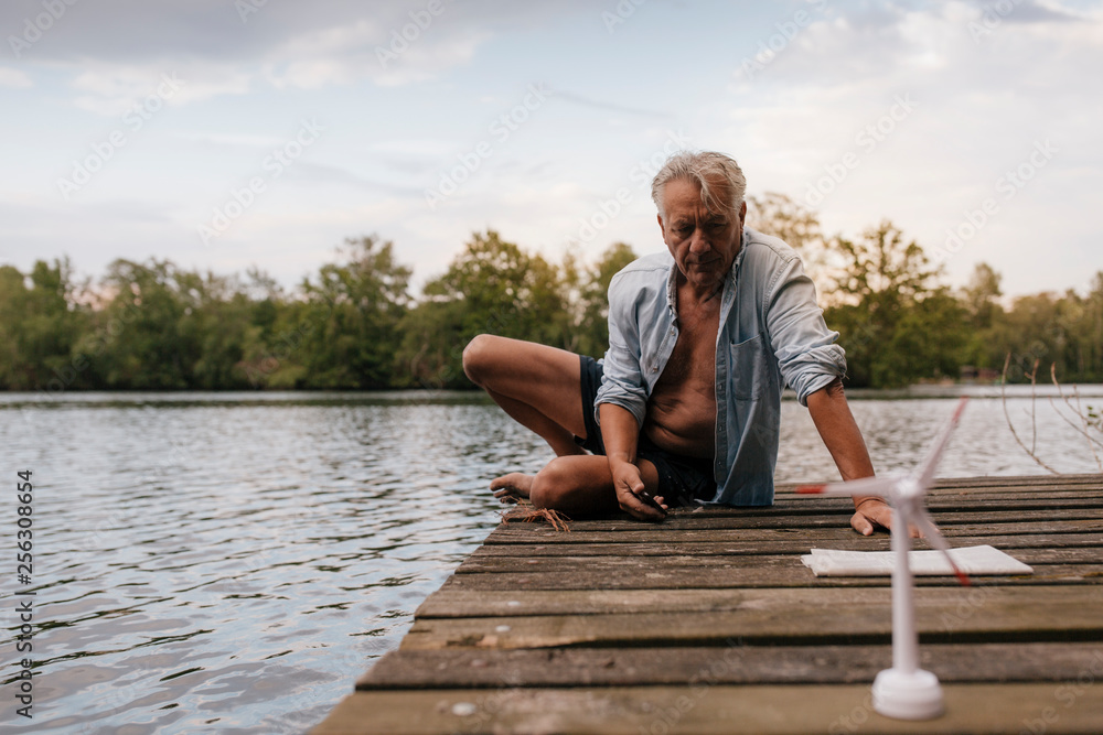Senior man sitting on jetty at a lake with small wind turbine model