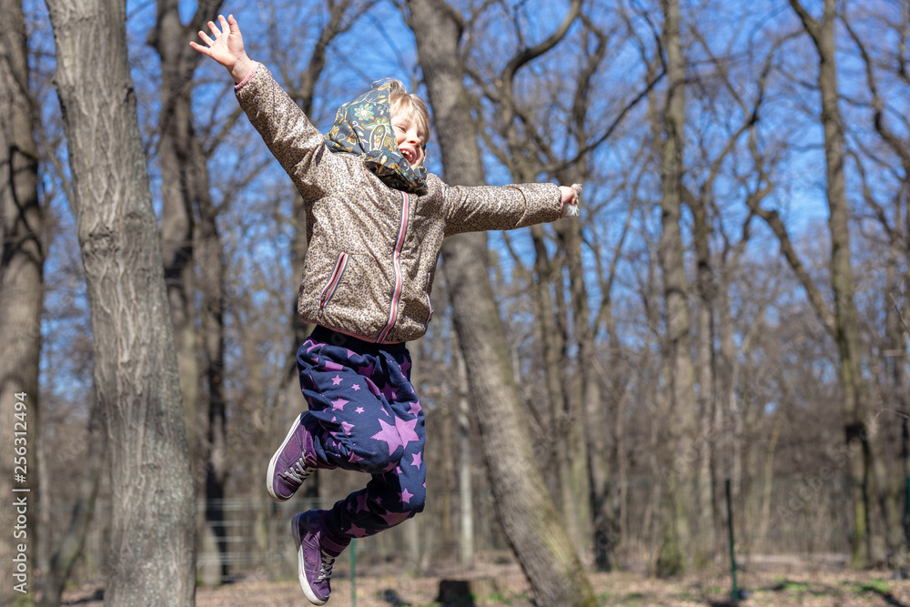 Cute little blond kid girl having fun outdoors. Child in casual sport wear and kerchief jumping high from tree stump in forest Cute little blond kid girl having fun outdoors. Child in casual sport wear and kerchief jumping high from tree stump in forest