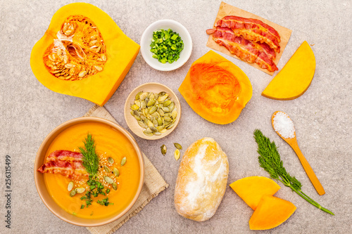 Homemade autumn hot pumpkin cream soup with smocked bacon and seeds. Raw pumpkin, bun, fresh chives, dill, salt in spoon, vintage linen cloth on stone background, top view, close up