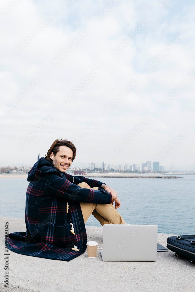 Spain, Barcelona, smiling man with laptop, briefcase and earphones sitting at the sea Spain, Barcelona, smiling man with laptop, briefcase and earphones sitting at the sea