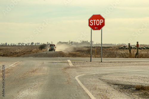 Country Roads - Stop sign with large bullet hole stands by crossroads with pickup truck with trailer driving toward it in a cloud of dust
