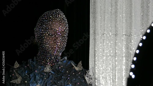 High-fashion. Model in the image of an alien girl in a black mask in sparkling rhinestones, sparkles, metal butterflies on a black and silver background posing in a model studio