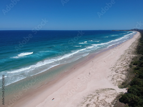 Drone view of an Australian beach. People walking on the white sand and the surf. Trees and grass on the sand dunes. Beautiful blue sky with a headland in the distance on the horizon.